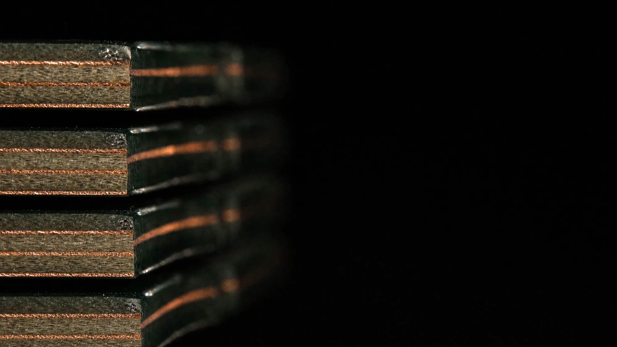 The cut edges of four circuit boards stacked with an offset, each showing a pale fibreglass core between thin bright copper layers, lit by a low raking light.